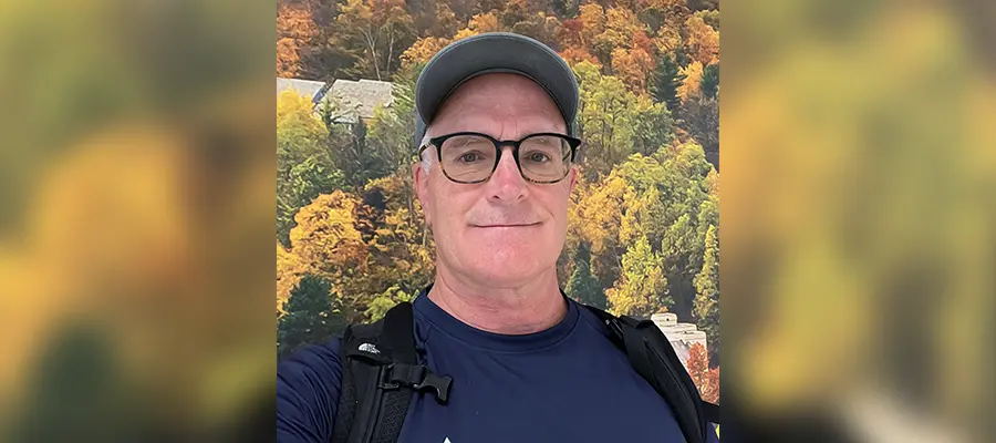 Raymond Jennings wearing black eyeglasses and a ball cap standing in front of foliage.