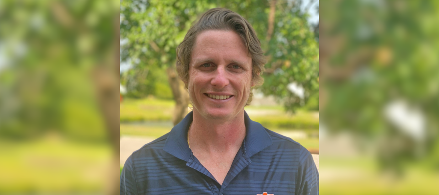 Joshua-Worsley-Headshot-BlurredBackground A smiling man wearing a blue collared shirt stands outdoors in a green park, with trees and grass behind him and a blurred extension of the background on both sides.
